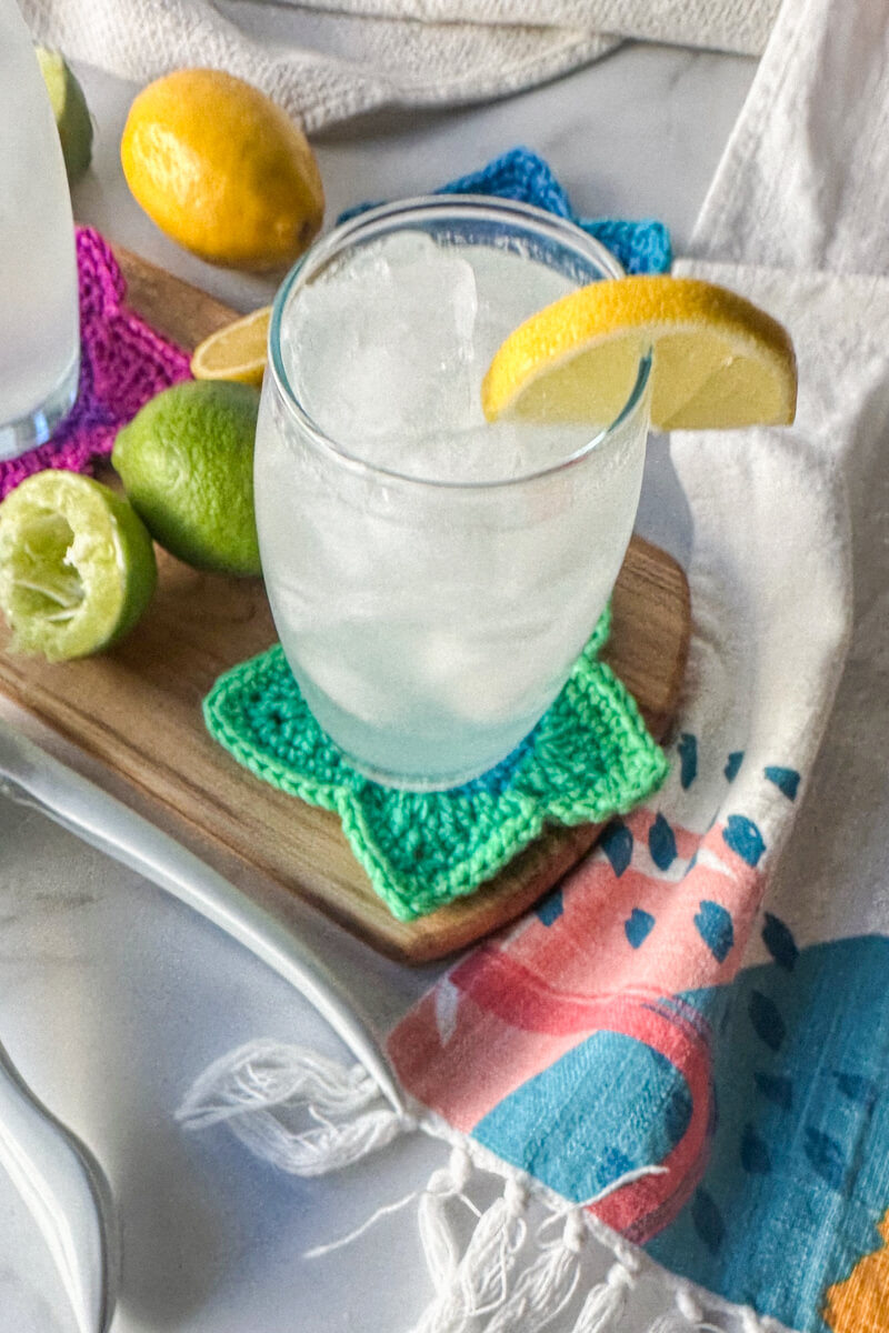 Overhead view of a clear glass filled with a low carb electrolyte drink, pale color, with ice and a lemon on the rim of the glass. Sitting on a wooden cutting board and crocheted green star coaster.