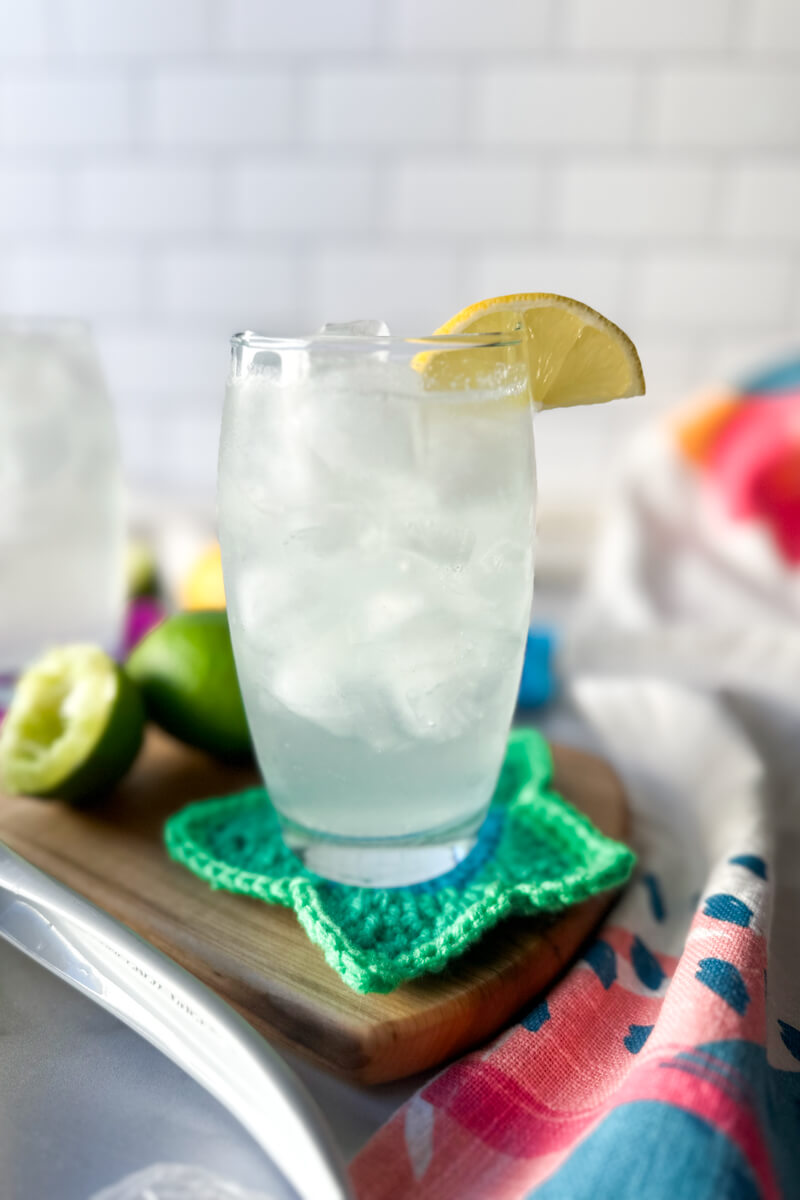 A clear glass filled with a low carb electrolyte drink, pale color, with ice and a lemon on the rim of the glass. Sitting on a wooden cutting board and crocheted green star coaster.