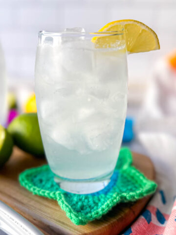 A clear glass filled with a low carb electrolyte drink, pale color, with ice and a lemon on the rim of the glass. Sitting on a wooden cutting board and crocheted green star coaster.