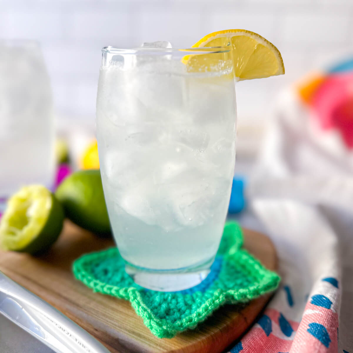 A clear glass filled with a low carb electrolyte drink, pale color, with ice and a lemon on the rim of the glass. Sitting on a wooden cutting board and crocheted green star coaster.
