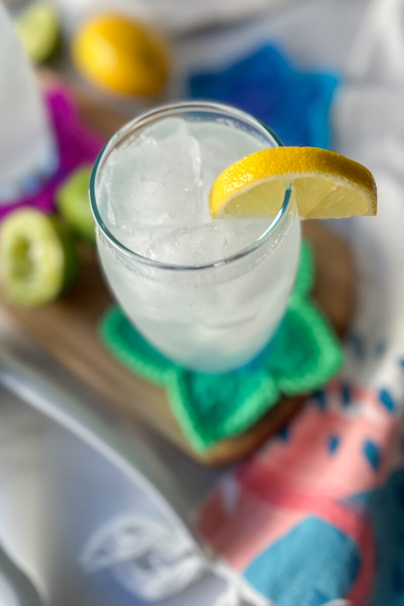 Overhead view of a clear glass filled with a low carb electrolyte drink, pale color, with ice and a lemon on the rim of the glass. Sitting on a wooden cutting board and crocheted green star coaster.