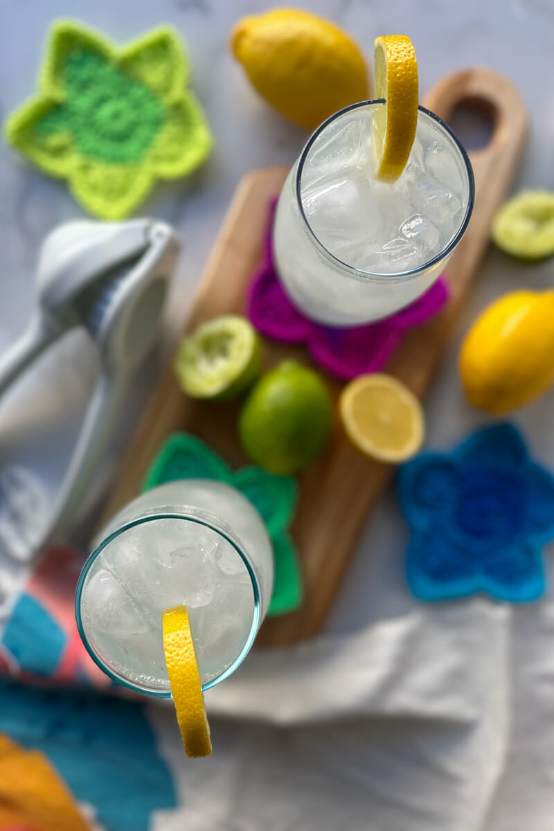 Overhead view of 2 clear glasses filled with a low carb electrolyte drink, pale color, with ice and a lemon on the rim of each glass. Sitting on a wooden cutting board and crocheted star coasters. Lemons, lime, and a juicer are lying next to them.