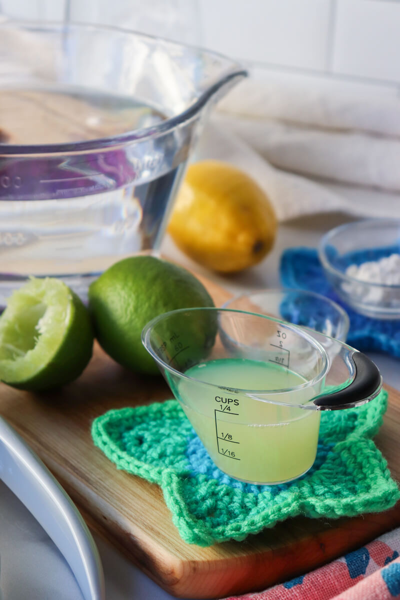 A glass measuring cup of water behind a smaller measuring cup of citrus juice, on wooden cutting board with lemons and limes.