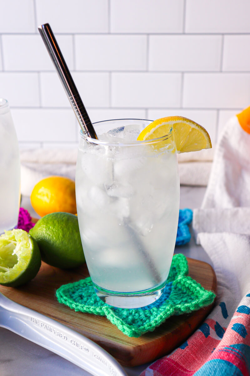 A clear glass filled with a low carb electrolyte drink, pale color, with ice and a lemon on the rim of the glass and steel straw. Sitting on a wooden cutting board and crocheted green star coaster.