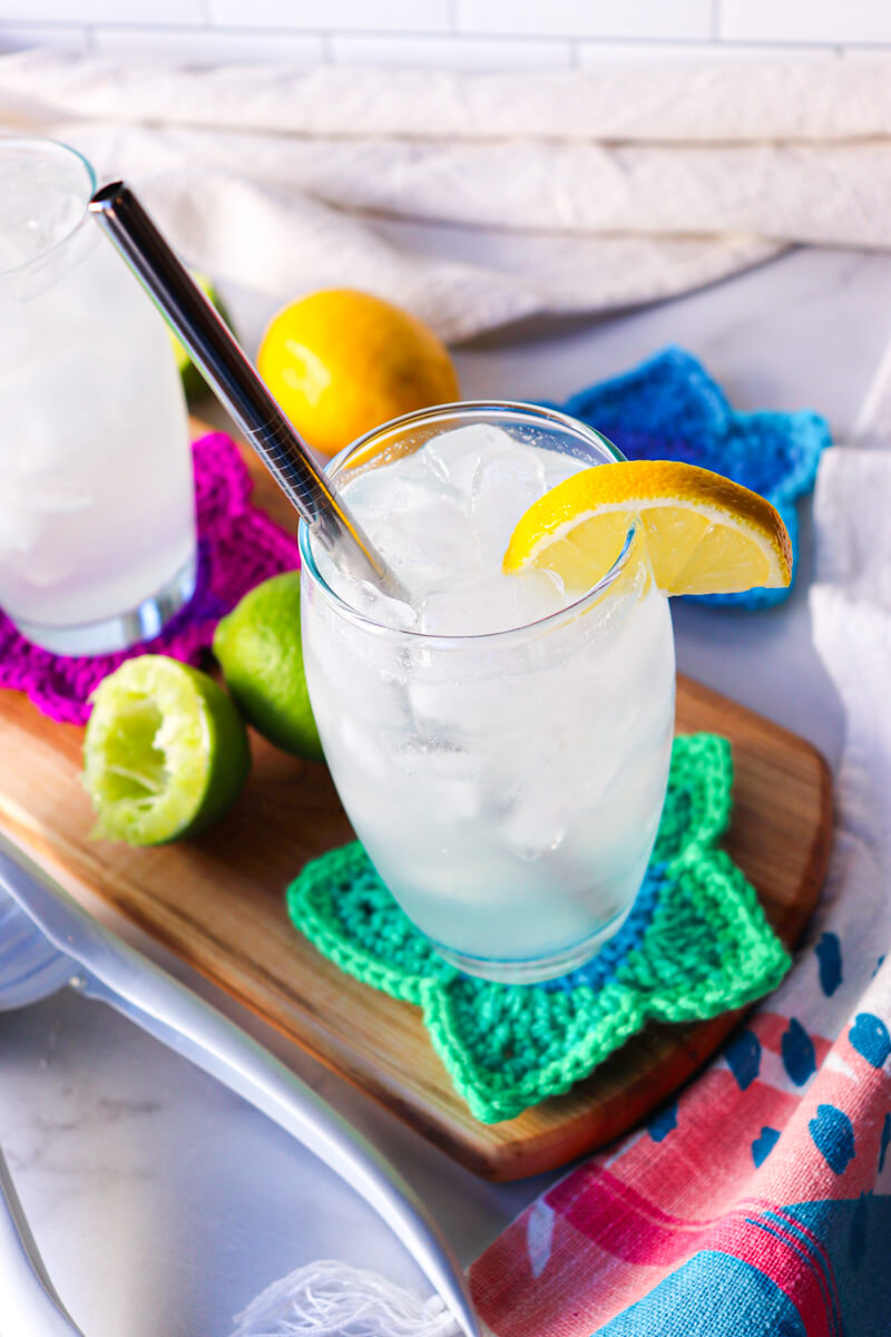 Overhead view of a clear glass filled with a low carb electrolyte drink, pale color, with ice and a lemon on the rim of the glass and steel straw. Sitting on a wooden cutting board and crocheted green star coaster.