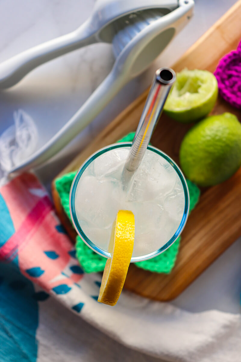 Overhead view of a clear glass filled with a low carb electrolyte drink, pale color, with ice and a lemon on the rim of the glass and steel straw. Sitting on a wooden cutting board and crocheted green star coaster.