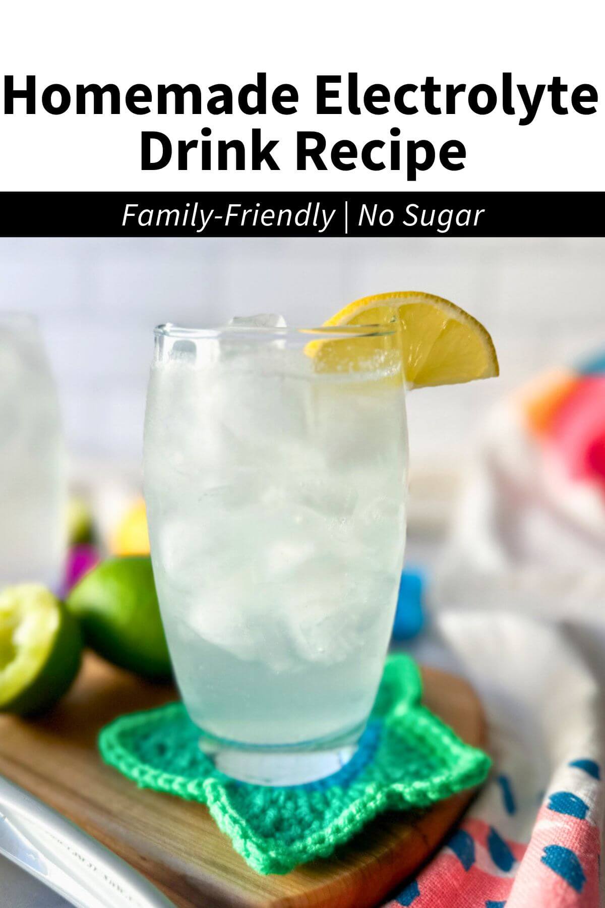 A clear glass filled with a low carb electrolyte drink, pale color, with ice and a lemon on the rim of the glass. Sitting on a wooden cutting board and crocheted green star coaster. Text overlay reads, "Homemade Electrolyte Drink Recipe, Family Friendly, no sugar."