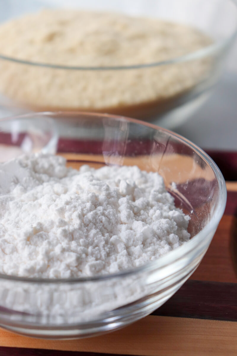 A glass bowl of white powdered sweetener on a striped wooden cutting board with another bowl in the background