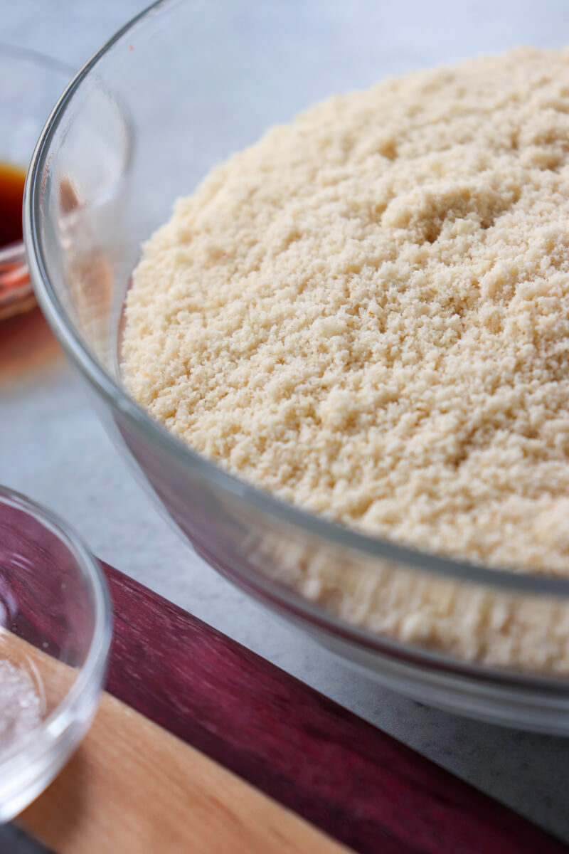 A glass bowl of finely ground almond flour next to a wooden striped cutting board