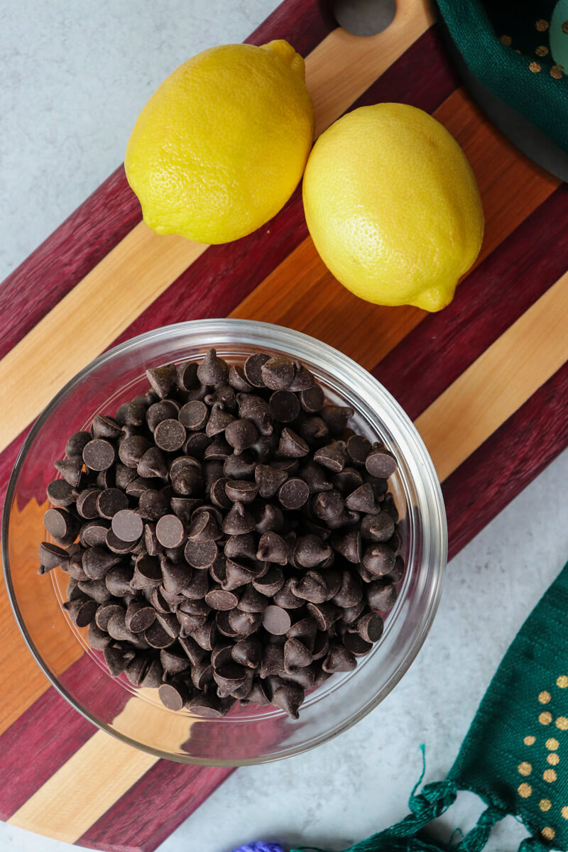 Overhead view of two lemons and a glass bowl of sugar free chocolate chips on a striped wooded cutting board
