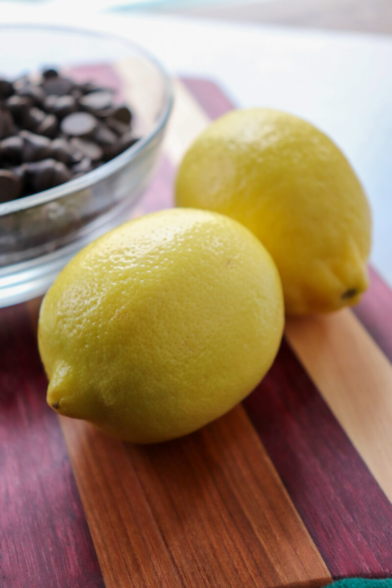 Two lemons and a glass bowl of sugar free chocolate chips on a striped wooded cutting board