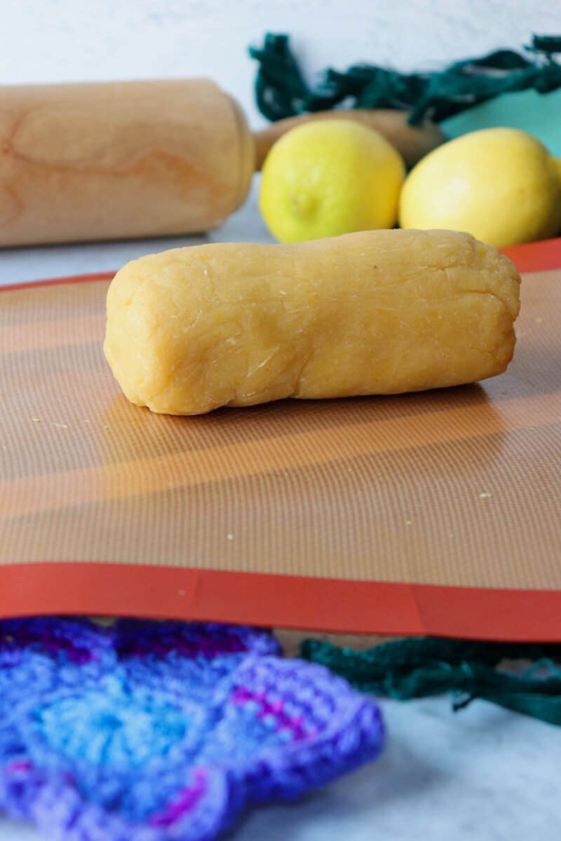 A chilled log of gluten free shortbread dough on a silicone mat with lemons and rolling pin in the background