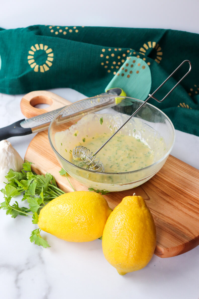 Front view of a glass bowl with a small whisk and the lemon dijon and parsley marinade on a wooden cutting board, with two lemons, parsley, and a zester next to it