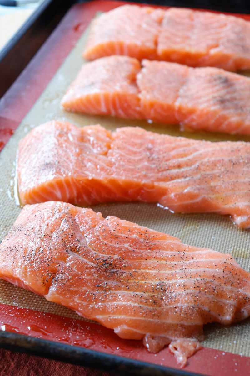 A pan of 4 raw sliced servings of salmon with olive oil, salt, and pepper, on a pan with a silicone baking mat