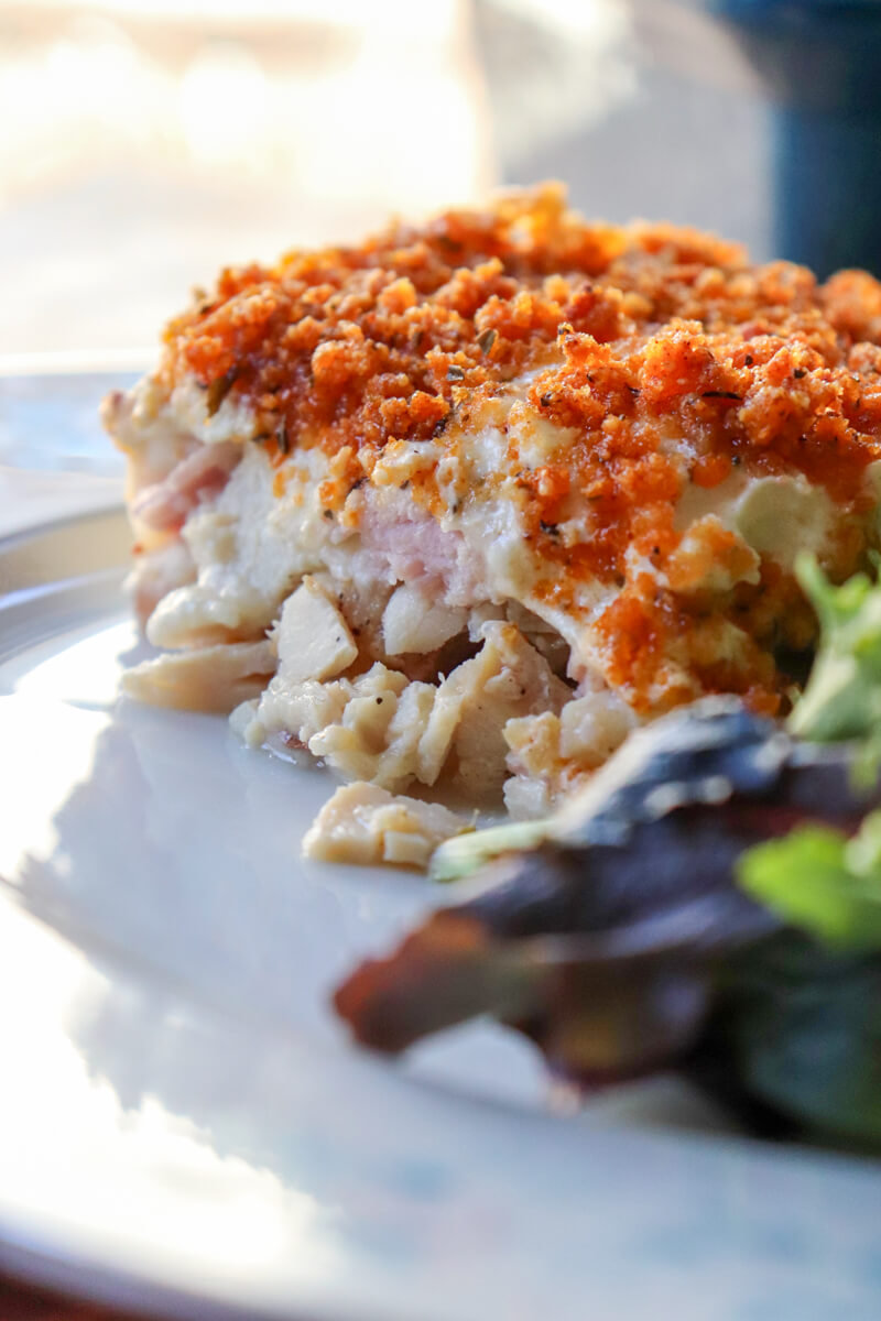 Close up of a slice of keto chicken cordon bleu casserole with crispy pork rind topping served on a plate with mixed greens salad and light from a window in the background.