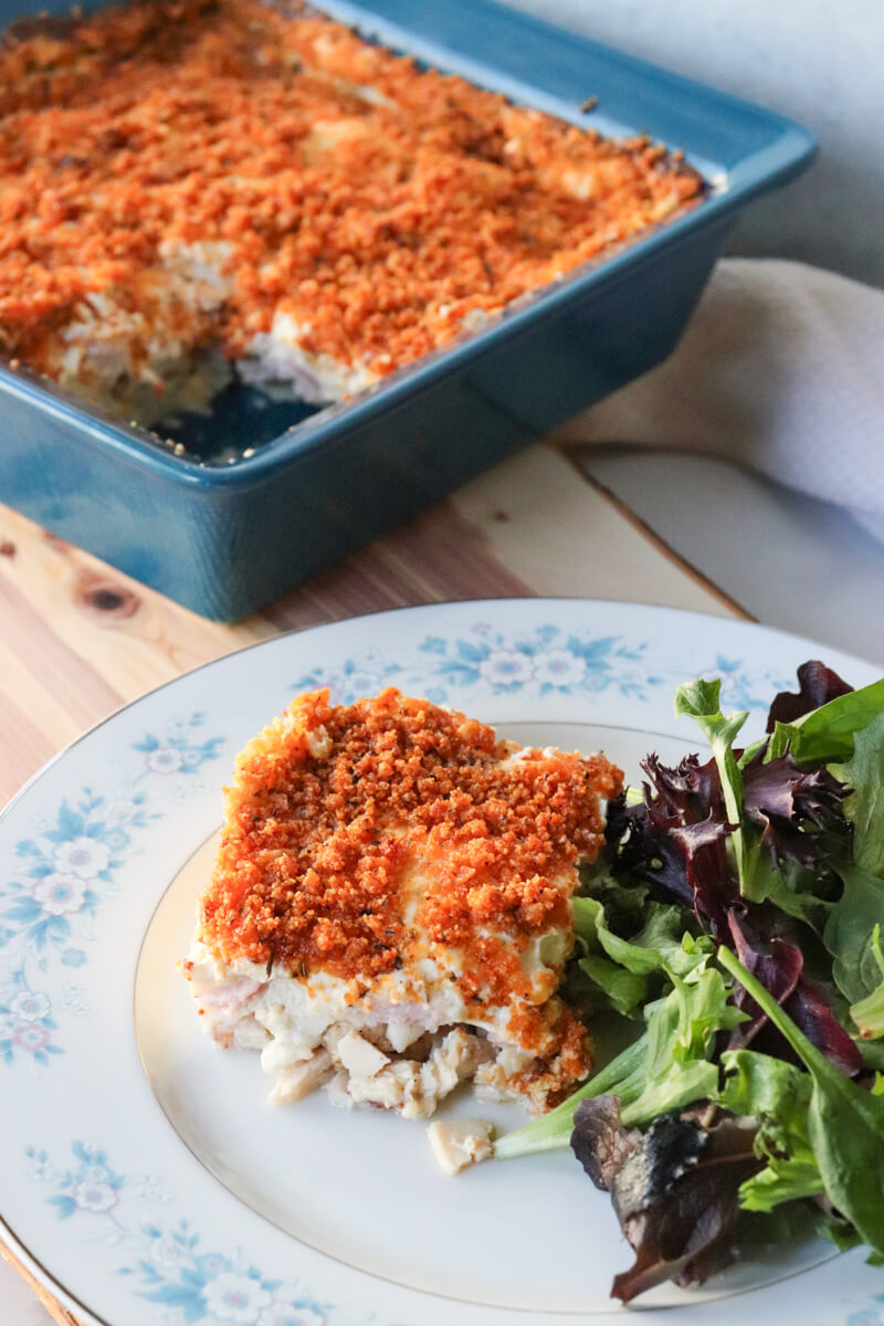 Slice of keto chicken cordon bleu casserole with crispy pork rind topping served on a plate with mixed greens salad in front of the blue serving dish with the rest of the casserole