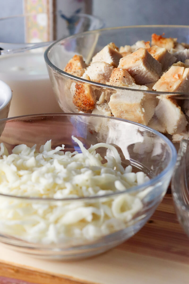 Small glass bowl of shredded mozzarella cheese in front of a glass bowl of diced chicken