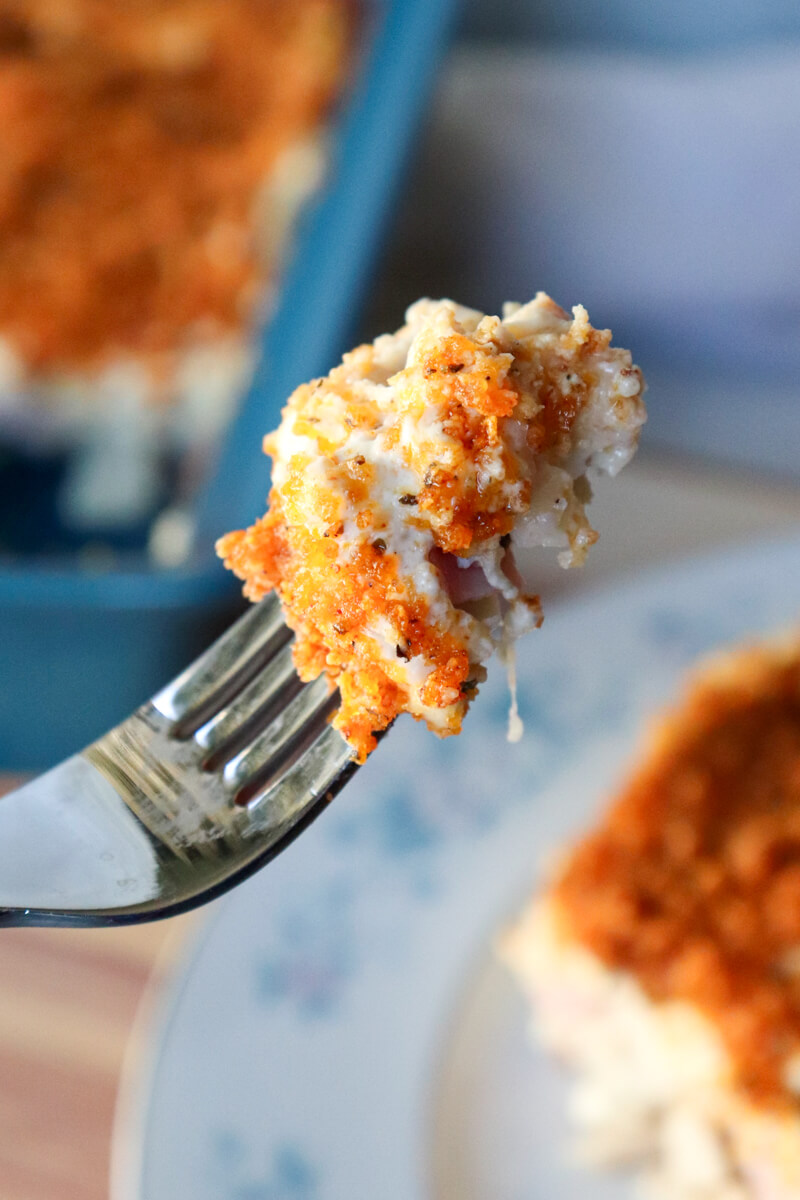A fork held up with a bite of keto chicken cordon bleu casserole with a serving and blue dish in the background.
