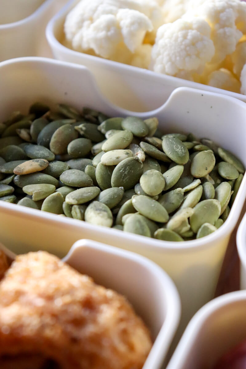 Close-up of keto snack containers with green pumpkin seeds and pork rinds arranged in a divided tray for a low carb movie snack setup