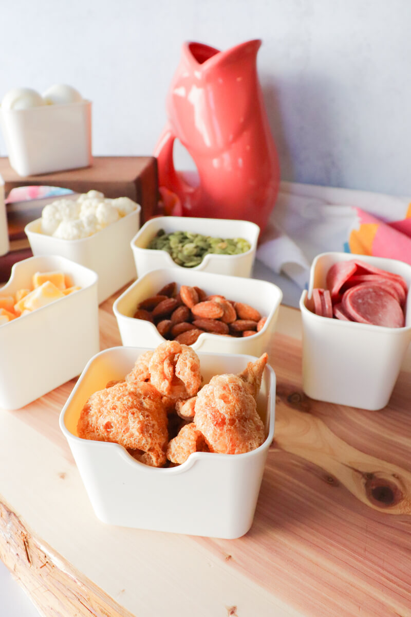 Keto movie snack setup on a wooden board with small bowls of cheese cubes, celery sticks, cauliflower, pumpkin seeds, almonds, pork rinds, salami, and hard boiled eggs arranged for a low carb snack spread