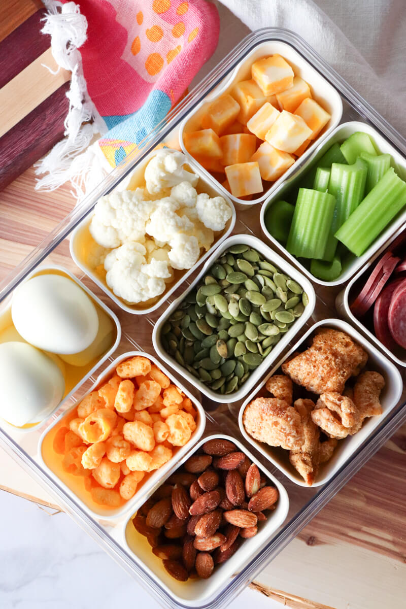 Low carb movie snacks in divided containers including almonds, cheese puffs, hard boiled eggs, pumpkin seeds, cauliflower, celery sticks, salami, and cheese cubes