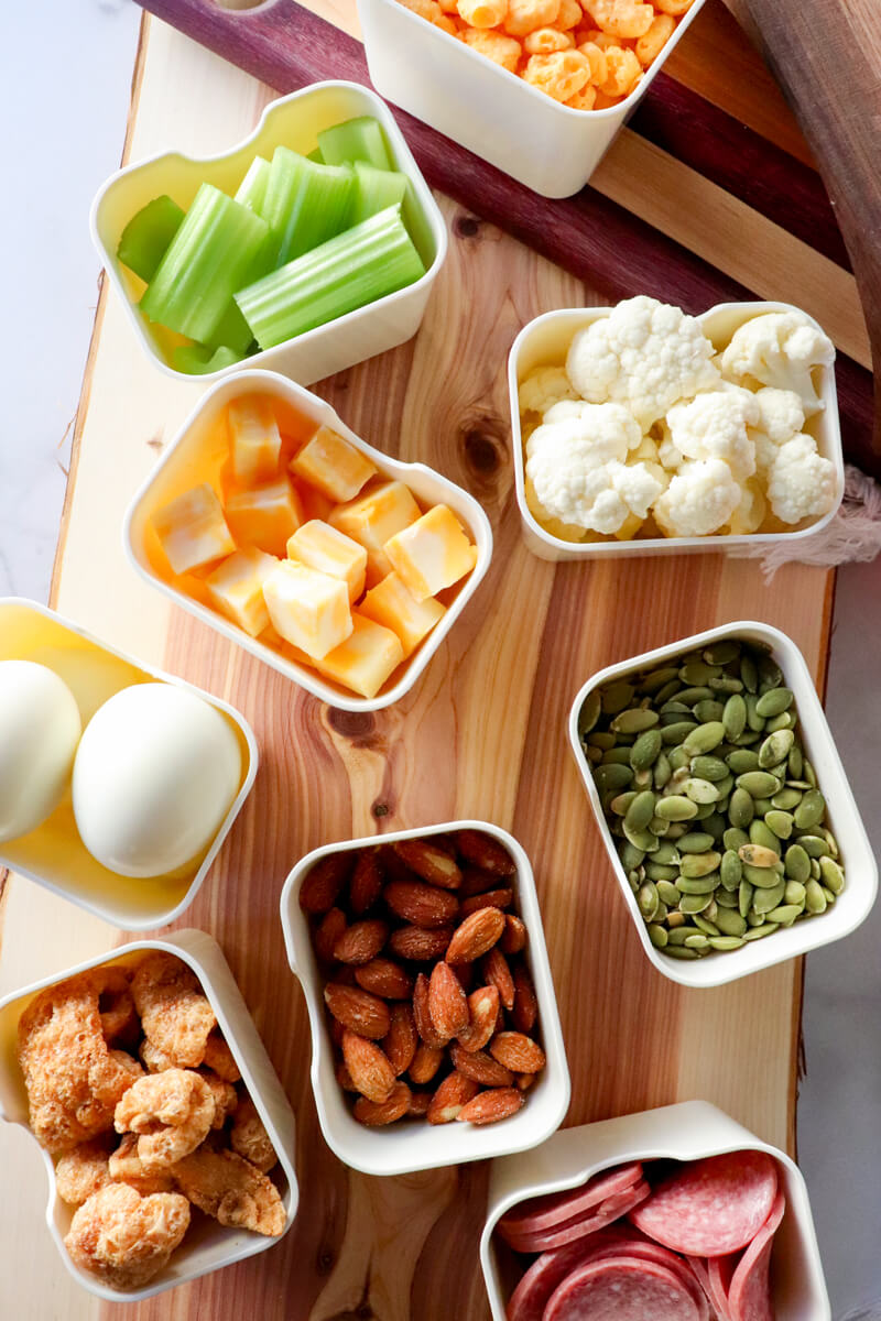 Overhead view of keto movie snacks arranged on a wooden board with small bowls of cheese cubes, celery sticks, cauliflower, pumpkin seeds, almonds, pork rinds, salami, and hard boiled eggs for a low carb snack spread