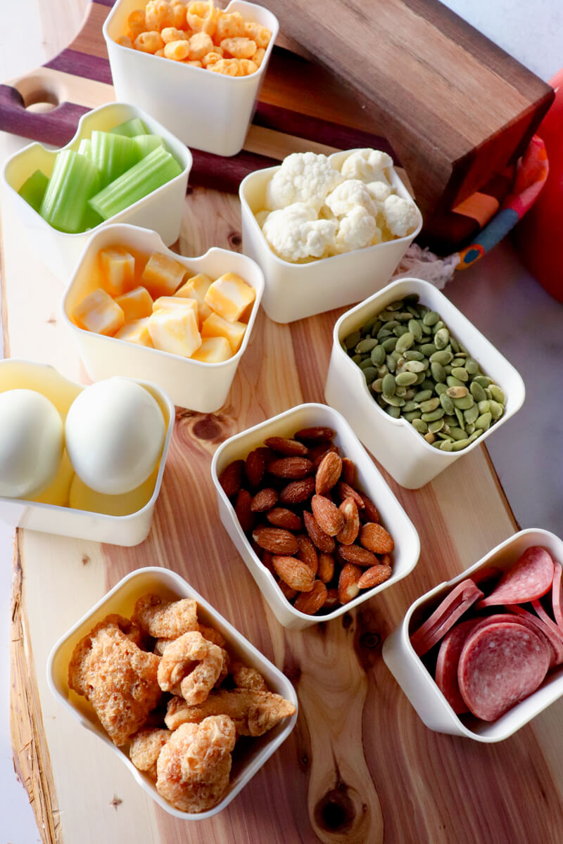 Keto movie snacks arranged on a wooden board with small bowls of cheese cubes, celery sticks, cauliflower, pumpkin seeds, almonds, pork rinds, salami, and hard boiled eggs for a low carb snack spread