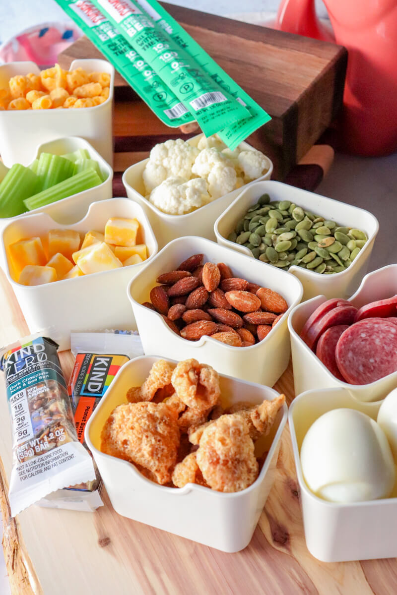 Keto snack spread on a wooden board with small bowls of cheese cubes, celery sticks, cauliflower, pumpkin seeds, almonds, pork rinds, salami, hard boiled eggs, and low carb snack bars arranged for a movie night