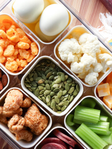Low carb movie snacks in divided containers including almonds, cheese puffs, hard boiled eggs, pumpkin seeds, cauliflower, celery sticks, salami, and cheese cubes