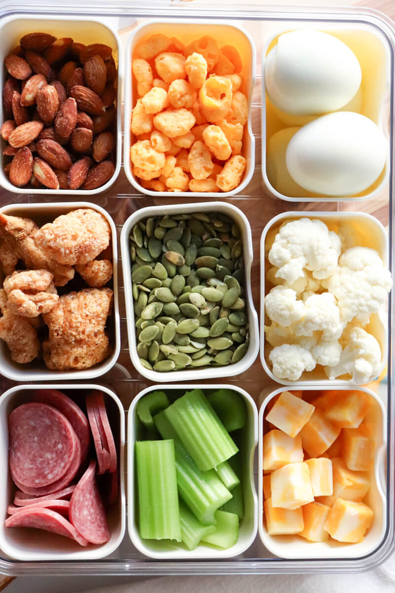 Low carb movie snacks in divided containers including almonds, cheese puffs, hard boiled eggs, pumpkin seeds, cauliflower, celery sticks, salami, and cheese cubes