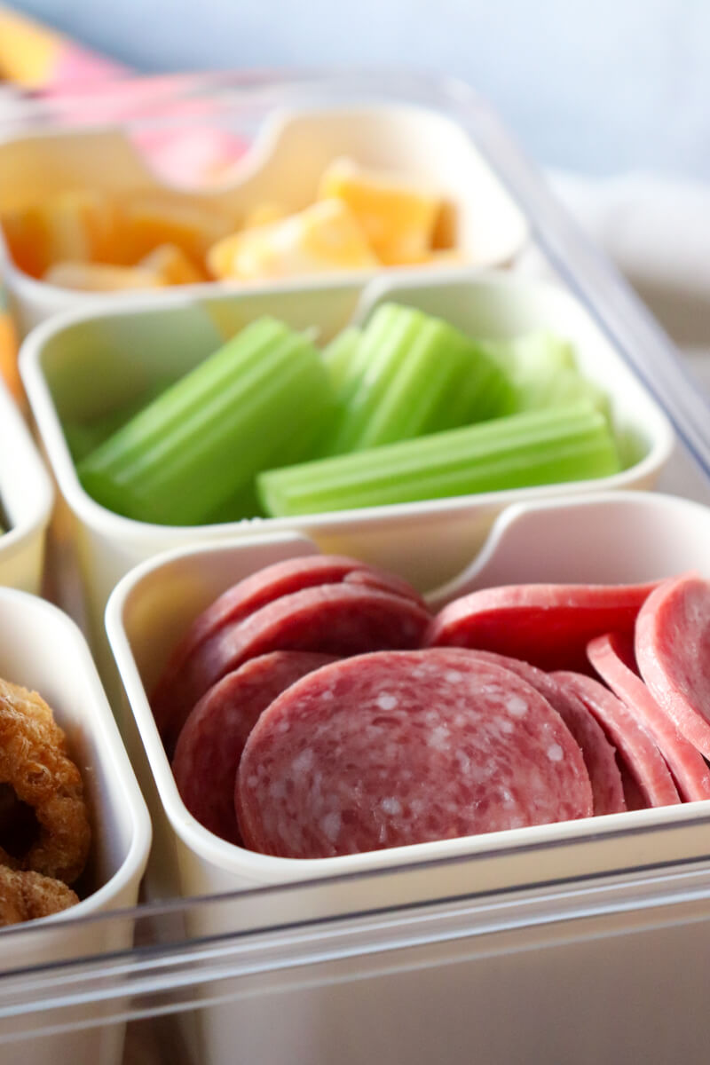 Close-up of keto snack containers with sliced salami, celery sticks, cheese cubes, and pork rinds arranged in a divided tray for a low carb movie snack setup