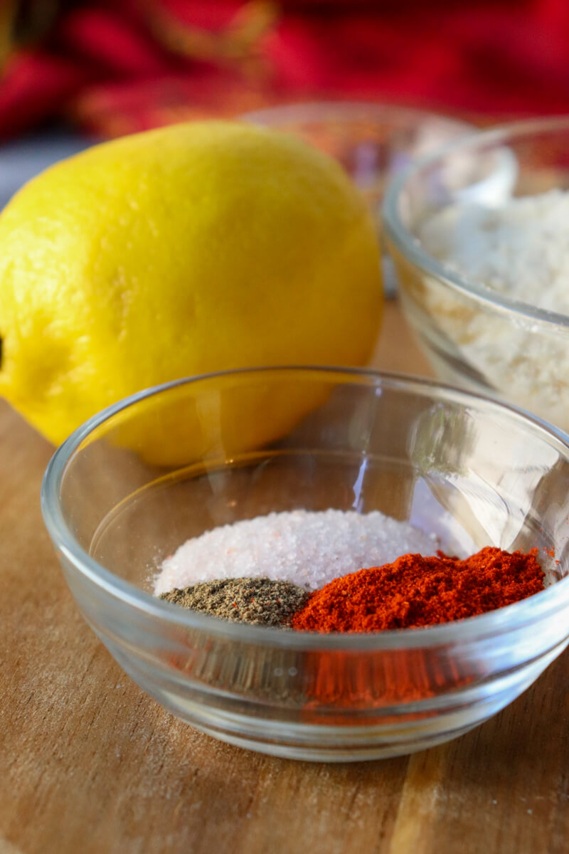 A glass bowl with salt, pepper, and paprika on a wooden board in front of a lemon