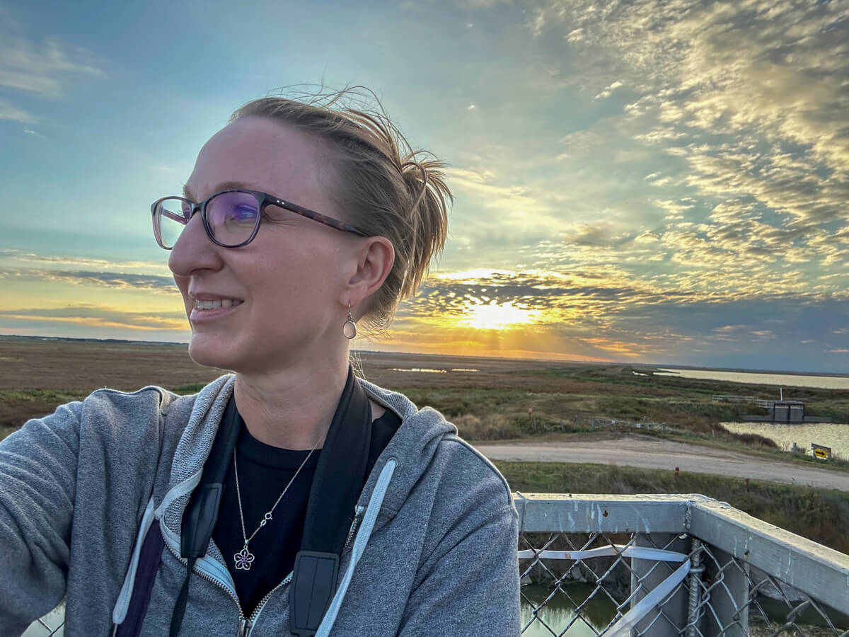 A women in glasses looking to the side in front of a sunset over the Kansas Wetlands.