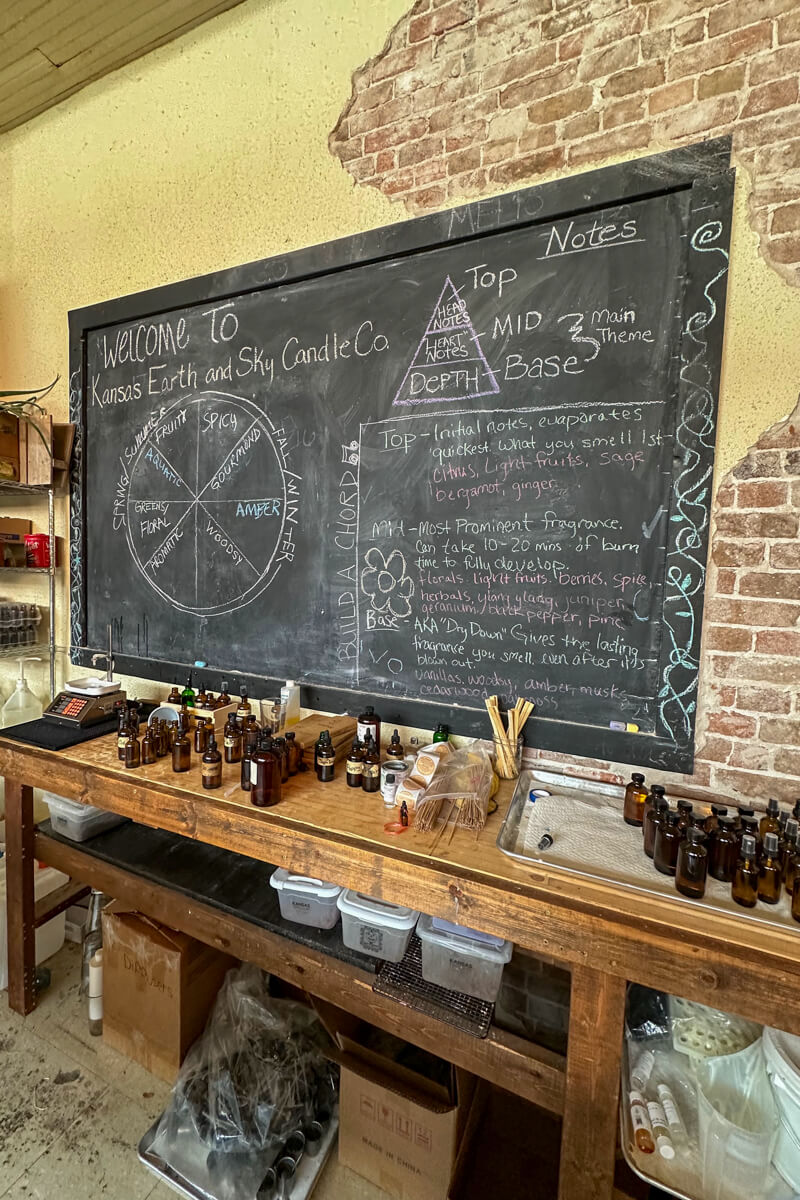 Chalkboard covered in writing over a wooden table with candle making supplies in the workshop area of Kansas Earth and Sky Candle Co.
