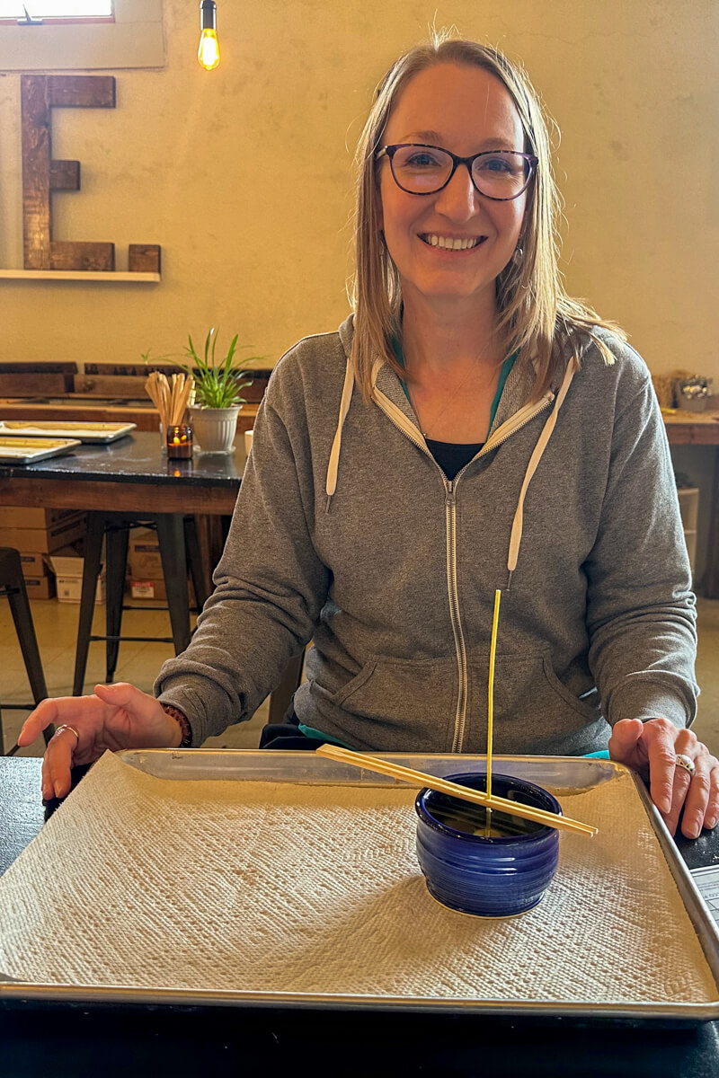 A woman in a gray zip sweatshirt and glasses sitting at a table with candle making supplies in the workshop area of Kansas Earth and Sky Candle Co.