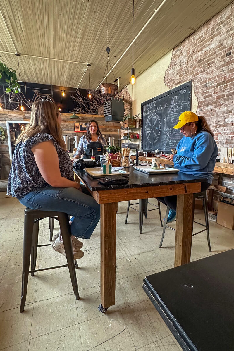 Two women and the teacher at a wooden table with candle making supplies in the workshop area of Kansas Earth and Sky Candle Co.