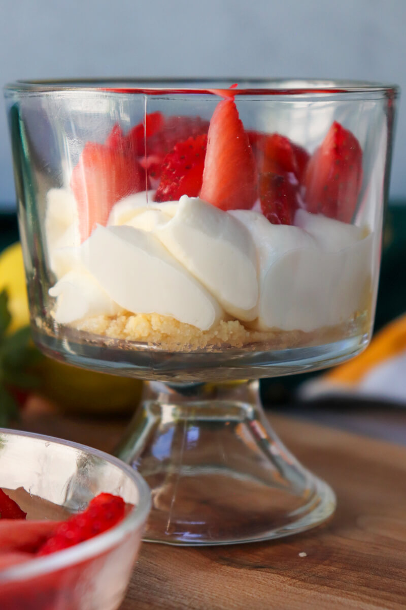 Front view of a low carb strawberry cheesecake in a small glass serving dish on pedestal. Bright layers of almond flour crust, cheesecake filling, and strawberry show through the glass.