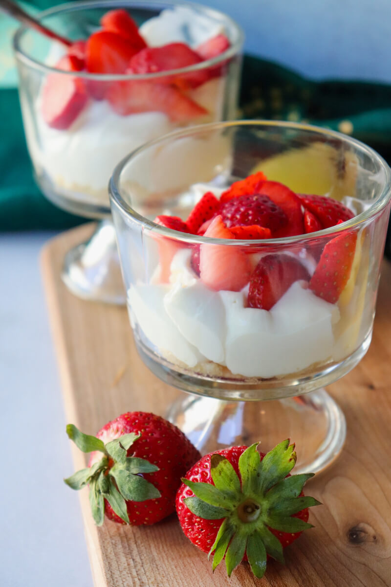 Two gluten-free no-bake strawberry cheesecake cups with whipped cheesecake filling and fresh strawberries served on a wooden board.