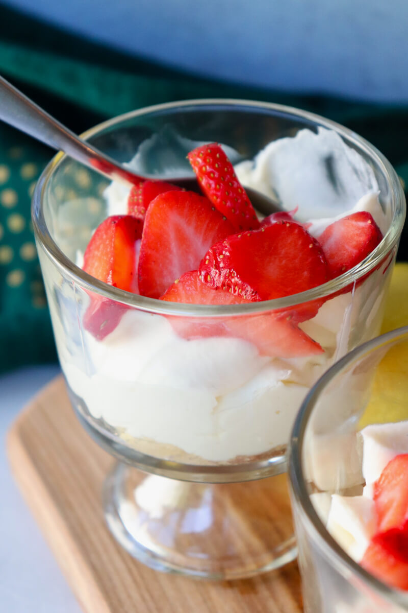 A strawberry cheesecake cup with whipped cheesecake filling, fresh strawberries, and a silver spoon, served on a wooden board.