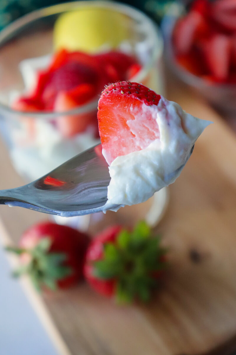 Spoonful of fluffy keto no-bake strawberry cheesecake with fresh strawberry slice in front of layered cheesecake dessert cups.