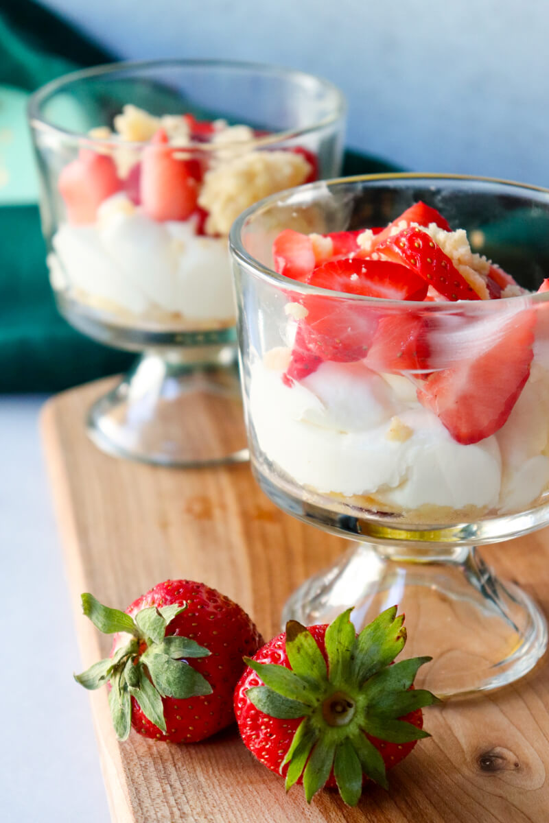 Front view of two gluten-free no-bake strawberry cheesecake cups with whipped cheesecake filling and fresh strawberries served on a wooden board.