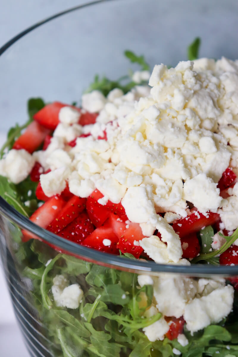 Overhead view of a glass serving bowl of strawberry arugula salad topped with feta cheese before being stirred