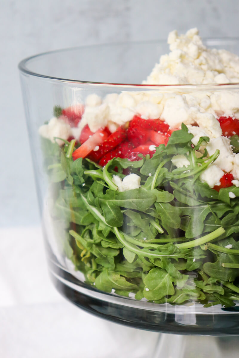 Glass serving dish with layers of strawberry arugula salad visible