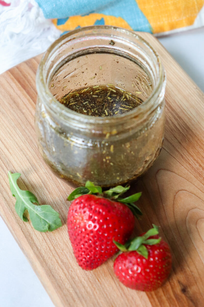 Overhead view of a small glass container of balsamic dressing with two strawberries on a wooden cutting board