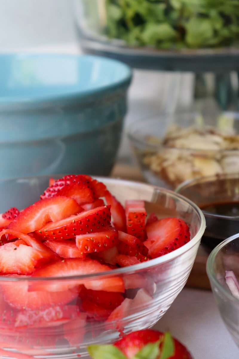 A clear glass bowl of sliced strawberries in front of a light blue ceramic bowl, and a glass bowl of sliced almonds