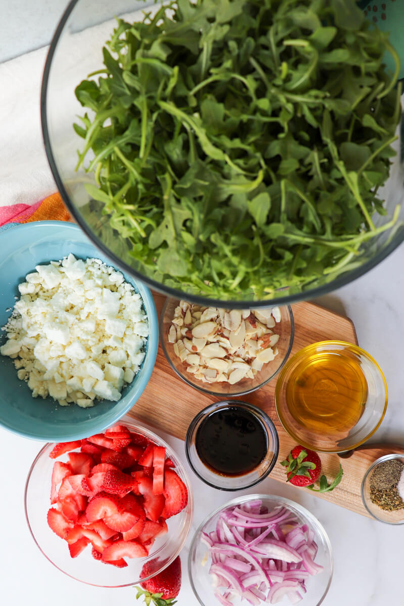 Overhead view of low carb strawberry arugula salad ingredients in bowls on a wooden cutting board