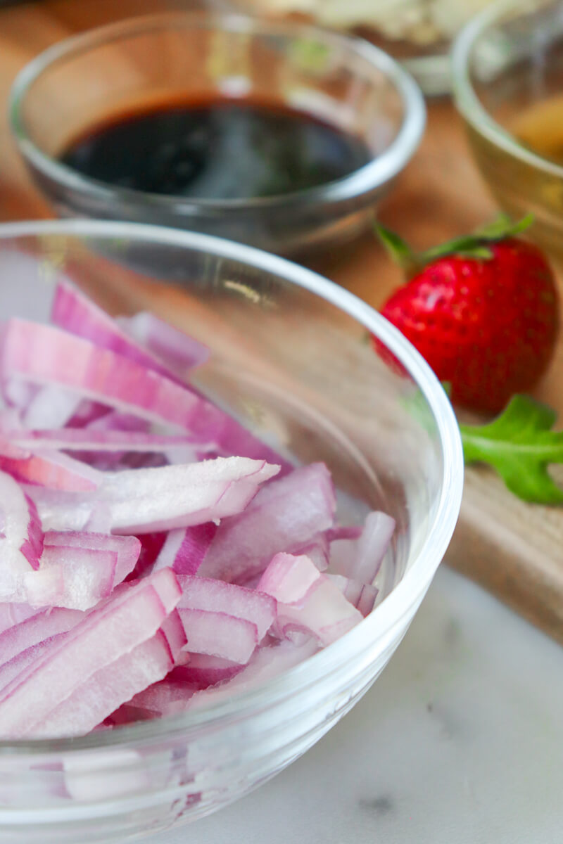 Glass bowls of red onion (in front), balsamic vinegar, and olive oil next to a strawberry