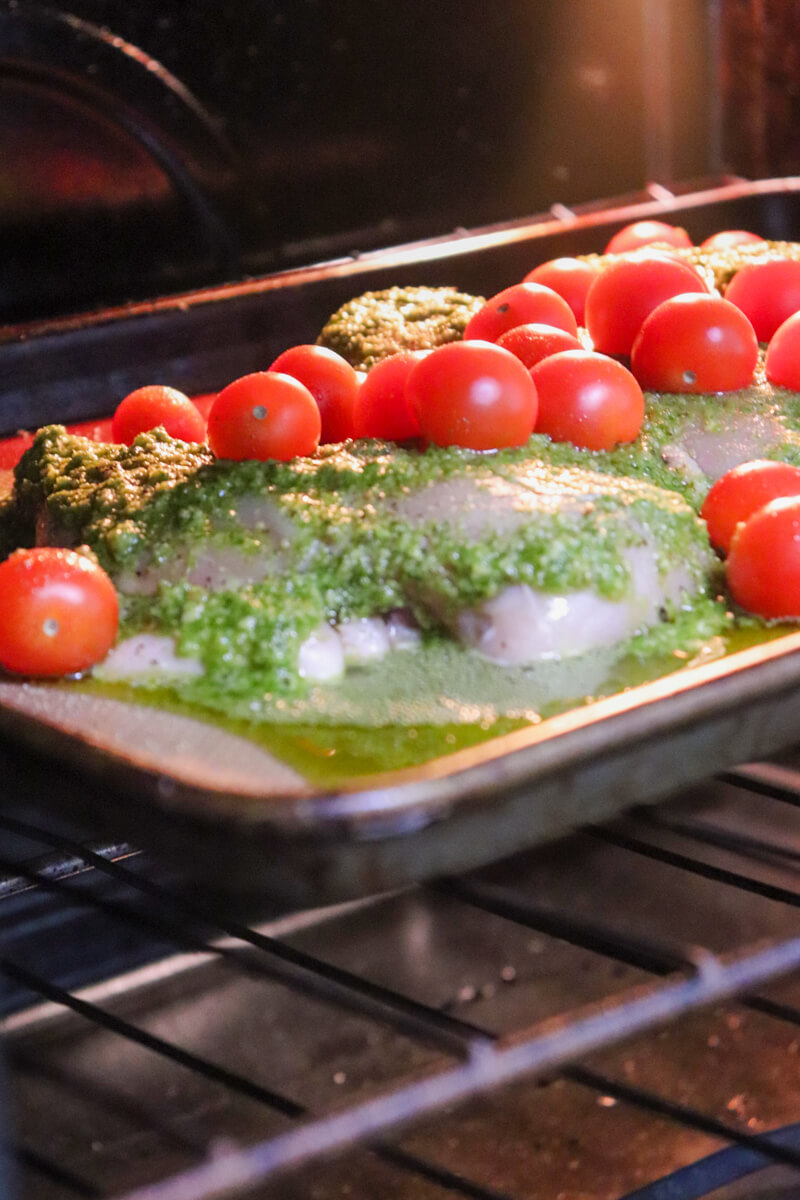 Raw prepped chicken with pesto and tomatoes on a pan in the oven