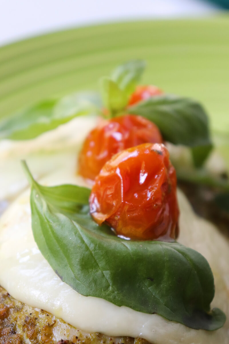 Close of of two tomatoes and basil leaves on low carb Pesto Chicken with Roasted Tomatoes & Mozzarella on a light green plate with arugula in the background