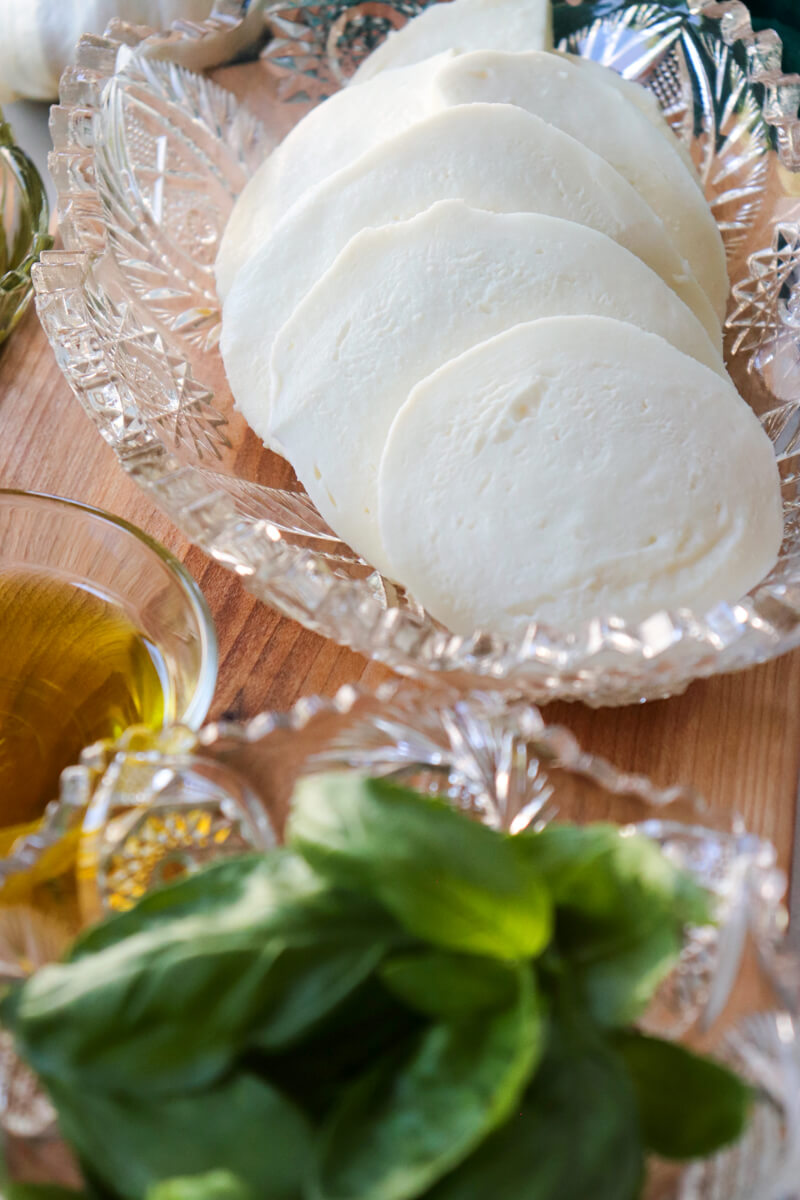 A decorative glass bowl of basil leaves in front of another with sliced fresh mozzarella and a small glass bowl of olive oil